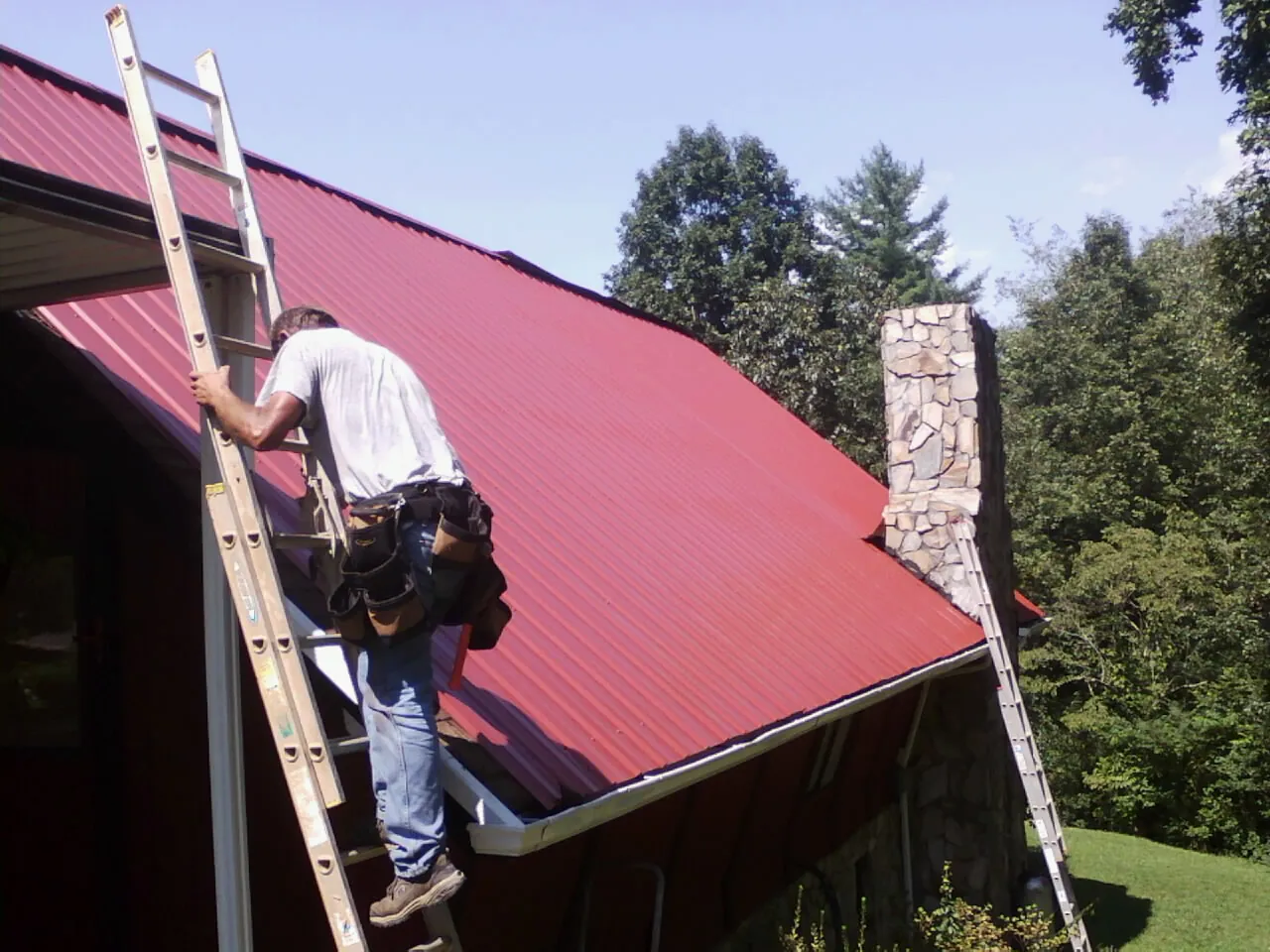 Roofer on a ladder beside a red metal roof and stone chimney — Soffit & Fascia Repair by Roofing Brothers in Malverne